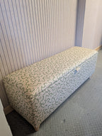 An upholstered storage bench with an Ian Sanderson fabric featuring geometric wavy pattern in pink and Green, set against a ribbed wall and textured gray workshop carpet.