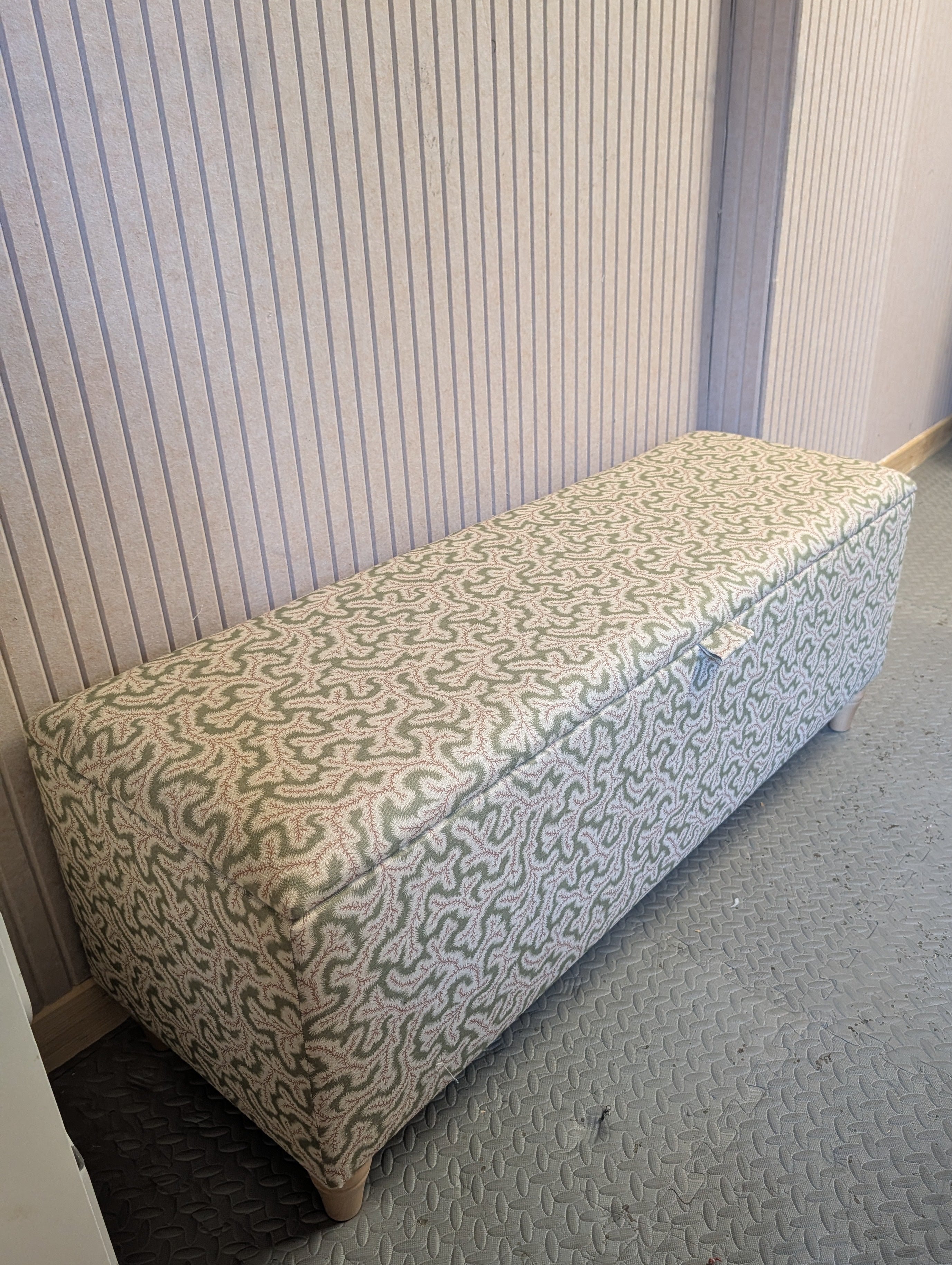 An upholstered storage bench with an Ian Sanderson fabric featuring geometric wavy pattern in pink and Green, set against a ribbed wall and textured gray workshop carpet.