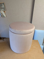 A cylindrical upholstered ottoman or stool with a round cushioned top, shown in a dusty blush pink velvet fabric. It is placed on a light brown wooden table against a beige ribbed wall.