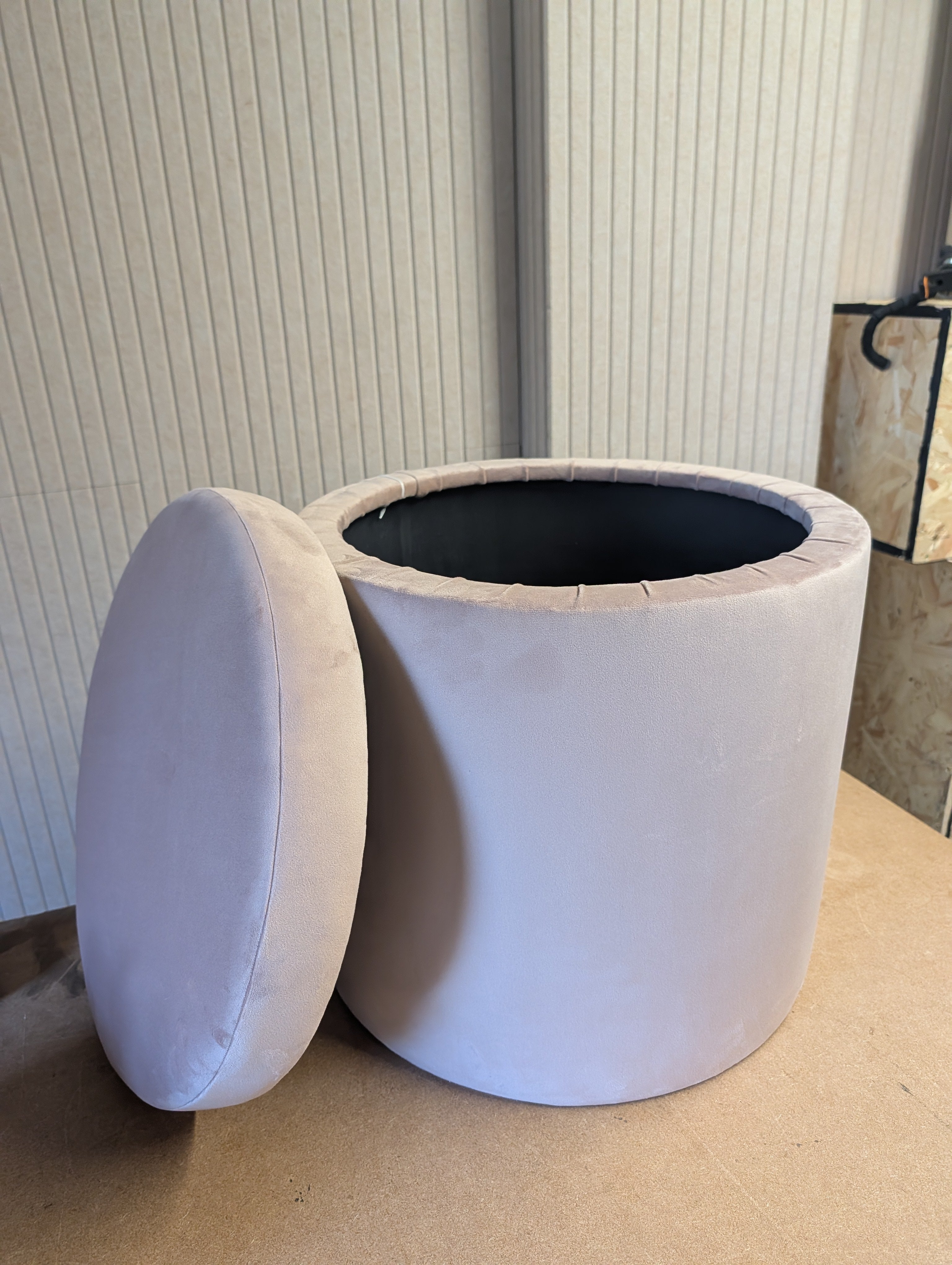 A cylindrical upholstered ottoman or stool with a matching cushioned lid leaning against it, shown on a tan floor in front of a ribbed wall.