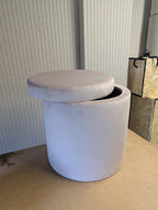 A cylindrical upholstered storage ottoman with a round cushioned lid, shown in a light pink petal fabric. The stool is photographed indoors on a tan floor against a ribbed panelled wall, with another furniture piece partially visible on the right.