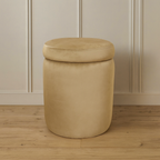Beige cylindrical ottoman with a lid on a wooden floor against a beige paneled wall.