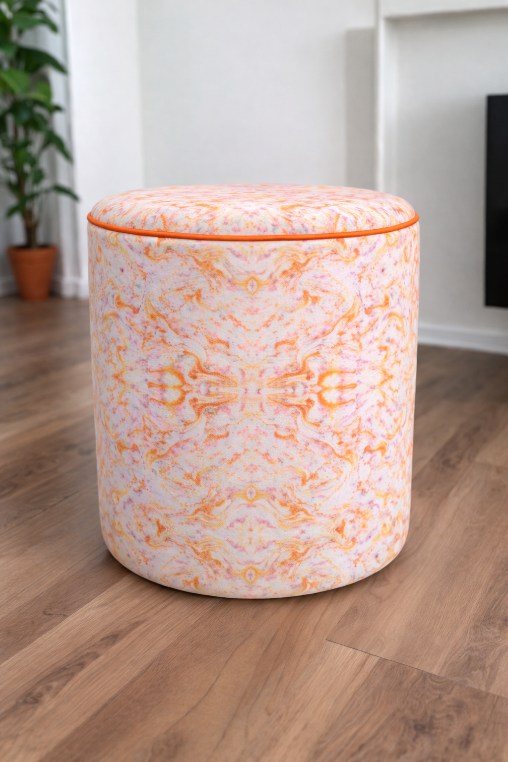 A cylindrical upholstered ottoman stool with a white base and an orange kaleidoscope retro pattern, set on a light wood floor in a bright room.