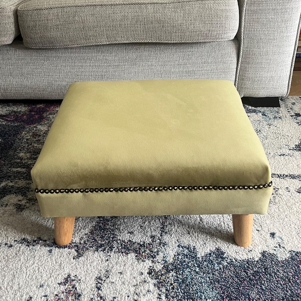 Short and low olive green footstool in plush velvet with bronze trim and angled wooden feet