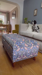 Opening of a William Morris Navy blue Strawberry Thief Velvet ottoman with a blanket being put inside and a black and white spaniel on the sofa behind watching. 